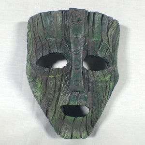 Puede incluir: Una máscara verde y marrón con una apariencia texturizada de madera. La máscara tiene aberturas para los ojos y la boca, una tira metálica vertical con remaches y un pequeño emblema en la parte superior. El diseño de la máscara recuerda a un accesorio de película clásico.
