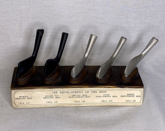 May include: A wooden display showcasing the evolution of the golf iron, with five miniature replicas of irons from different eras. The display includes labels indicating the type of iron and the approximate year of its introduction, starting with the "Spurtoe Wrought Iron" from circa 1620 and ending with the "Mashie Drop-Forged Iron" from circa 1905.