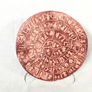 May include: A round terracotta plate with a spiral design of symbols and figures. The plate is a reddish-brown color and has a white background.