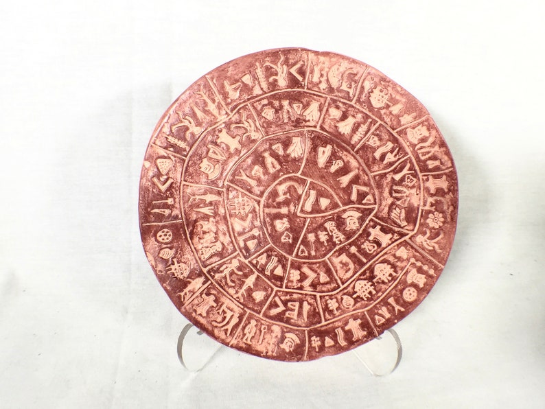 May include: A round terracotta plate with a spiral design of symbols and figures. The plate is a reddish-brown color and has a white background.