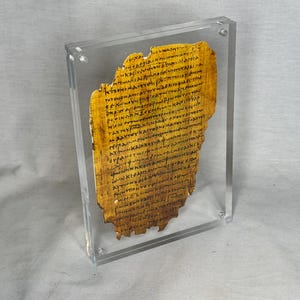Puede incluir: Un fragmento de papiro antiguo enmarcado. El papiro es de color marrón claro con texto negro. El fragmento está encerrado en un marco de acrílico transparente, mostrando el documento envejecido. El texto está escrito en una escritura.