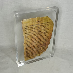Puede incluir: Un fragmento de papiro antiguo enmarcado con texto escrito a mano en tinta negra. El papiro está amarillento por la edad y tiene un borde ligeramente desgarrado. El marco es de acrílico transparente.