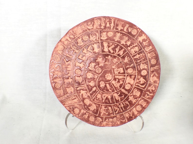 May include: A terracotta circular plate with a spiral design of various symbols and figures. The plate is on a white background.
