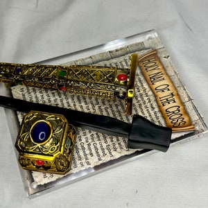 Könnte beinhalten: Eine goldfarbene Nachbildung eines Nagels vom Kreuz, mit aufwendigen Details und roten und grünen Edelsteinen. Die Nachbildung wird auf einem durchsichtigen Acryl-Ständer mit einer gedruckten Beschreibung des Heiligen Nagels vom Kreuz präsentiert.