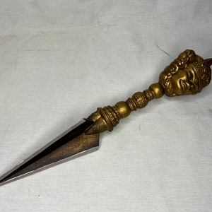 Puede incluir: Una daga ceremonial de color dorado con un mango detallado y ornamentado que presenta un rostro humano estilizado. La hoja es afilada y puntiaguda.