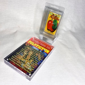 Puede incluir: Un expositor de acrílico transparente con un paquete de cigarrillos "Red Apple" rojo y amarillo en su interior. El expositor está al lado de otro expositor de acrílico transparente con un póster de la película Pulp Fiction en su interior. El póster tiene un fondo negro con texto e imágenes blancas.