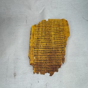 Puede incluir: Un fragmento de papiro antiguo con escritura en tinta negra descolorida. El papiro tiene un aspecto amarillento y envejecido con bordes irregulares, lo que sugiere su edad e importancia histórica. El texto está densamente escrito.