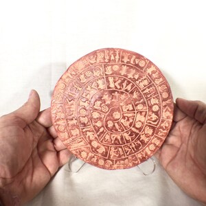 May include: A round, terracotta-colored ceramic plate with a spiral design of various symbols and figures. The plate is on a white background.