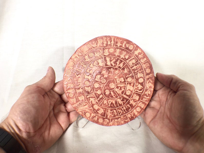 May include: A round, terracotta-colored ceramic plate with a spiral design of various symbols and figures. The plate is on a white background.