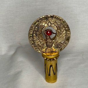 Può includere: Tappi di bottiglia dorati con un design circolare che presenta una creatura alata con un gioiello rosso nell'occhio. Il design è circondato da geroglifici.