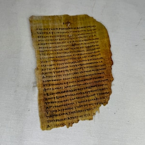 Puede incluir: Un fragmento de un rollo de papiro amarillento con escritura en tinta negra en griego antiguo. El texto es parcialmente visible e incluye las palabras "Χριστιανων" y "εγγραφη."