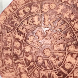 May include: A circular terracotta plate with a spiral design of various symbols and patterns. The plate is decorated with a repeating pattern of small circles, stars, and other geometric shapes.