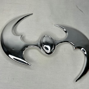 May include: A silver-coloured metal batarang, a throwing weapon shaped like a bat, with a rounded centre and two pointed wings.