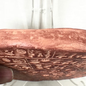 May include: A terracotta-colored circular disc with intricate carvings and designs. The disc is slightly raised in the center and has a smooth, slightly textured surface.