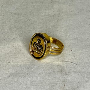 May include: A gold ring with an oval face displaying a detailed, stylized design. The ring has a triple-band design and a polished finish. The design on the face appears to be a figure with a crown.