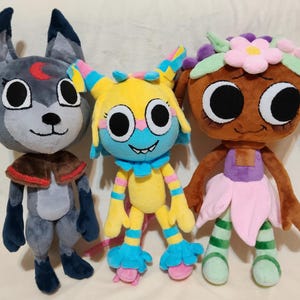 May include: Three plush toys standing side by side. The left is a gray wolf-like creature with a red crescent moon on its forehead. The center is a yellow, blue, and pink creature with large eyes. The right is a brown doll with a flower crown.