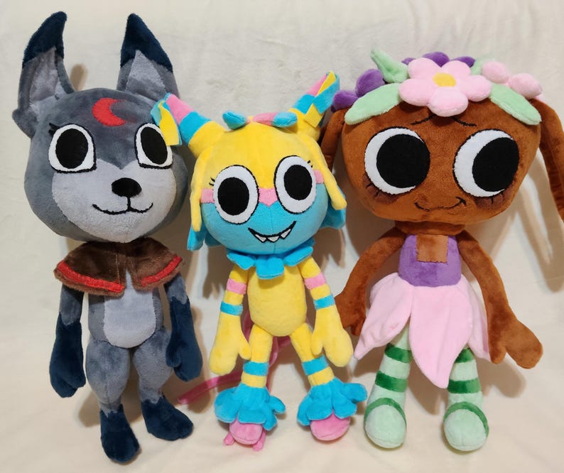 May include: Three plush toys standing side by side. The left is a gray wolf-like creature with a red crescent moon on its forehead. The center is a yellow, blue, and pink creature with large eyes. The right is a brown doll with a flower crown.