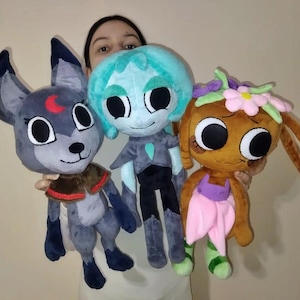 May include: Three plush toys featuring large black eyes. The left toy is a gray wolf-like creature with a red crescent moon on its forehead. The center toy is a teal-haired figure in a dark outfit. The right toy is brown with pink flowers.