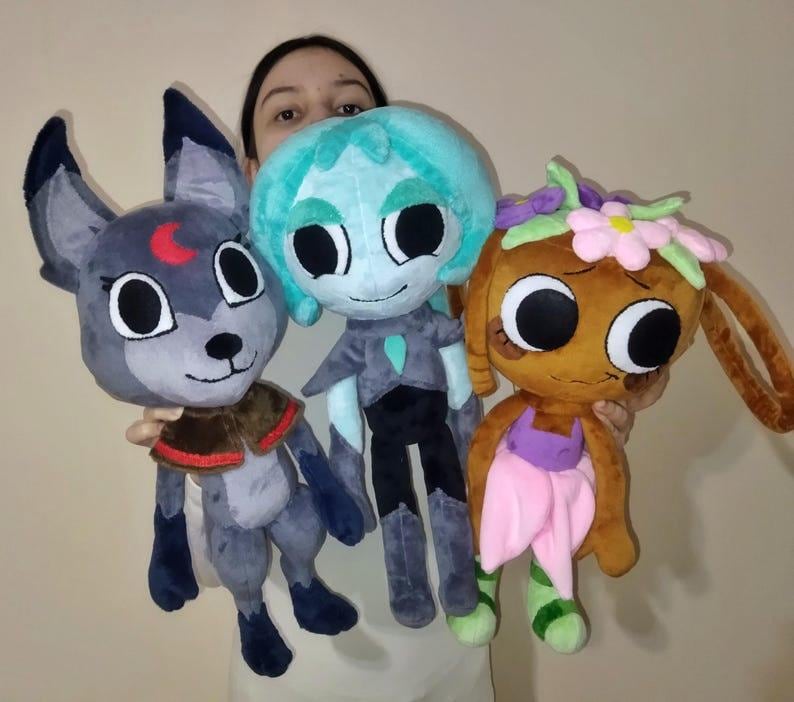 May include: Three plush toys featuring large black eyes. The left toy is a gray wolf-like creature with a red crescent moon on its forehead. The center toy is a teal-haired figure in a dark outfit. The right toy is brown with pink flowers.