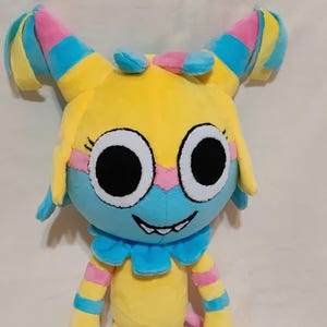 May include: A plush toy featuring a yellow head with large white eyes and black pupils, a blue lower face, and a wide grin with visible teeth. The toy has two tall, pointed ears with pink, blue, and yellow stripes, and a blue ruffled collar.