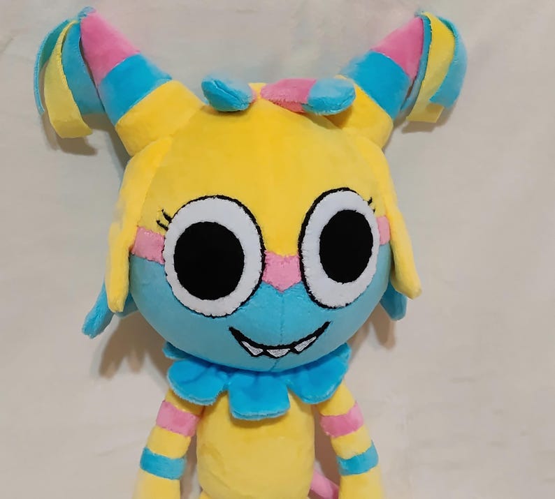 May include: A plush toy featuring a yellow head with large white eyes and black pupils, a blue lower face, and a wide grin with visible teeth. The toy has two tall, pointed ears with pink, blue, and yellow stripes, and a blue ruffled collar.
