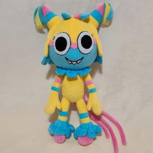 May include: A plush toy featuring a yellow head with large white and black eyes, a blue face mask, and a wide grin with visible teeth. The body is yellow with blue and pink striped limbs, blue ruffled cuffs at the neck, wrists, and ankles.