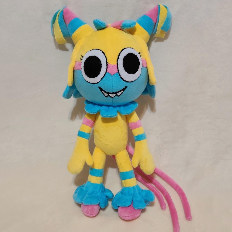 May include: A plush toy featuring a yellow head with large white and black eyes, a blue face mask, and a wide grin with visible teeth. The body is yellow with blue and pink striped limbs, blue ruffled cuffs at the neck, wrists, and ankles.