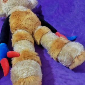 Peut inclure: Une poup&eacute;e en peluche au design fantaisiste. La poup&eacute;e a une t&ecirc;te beige avec des yeux noirs et une bouche souriante. Le corps est beige et orange, avec des bras et des jambes noirs. Les pieds sont beiges et orange, avec des accents rouges et bleus. La poup&eacute;e est pos&eacute;e sur une surface violette.