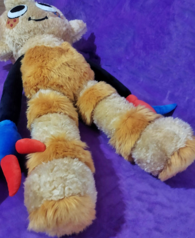 Peut inclure: Une poup&eacute;e en peluche au design fantaisiste. La poup&eacute;e a une t&ecirc;te beige avec des yeux noirs et une bouche souriante. Le corps est beige et orange, avec des bras et des jambes noirs. Les pieds sont beiges et orange, avec des accents rouges et bleus. La poup&eacute;e est pos&eacute;e sur une surface violette.