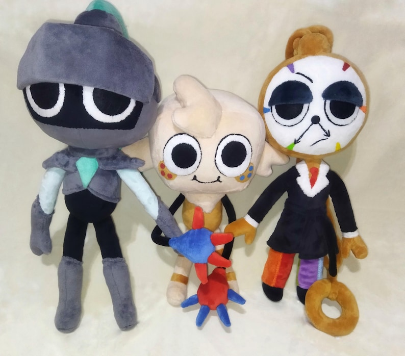 Peut inclure: Trois poup&eacute;es en peluche aux traits de dessins anim&eacute;s. La premi&egrave;re est grise avec un casque. La seconde est beige et tient un jouet en forme d'&eacute;toile bleu et rouge. La troisi&egrave;me porte un costume noir et a un visage d'horloge.