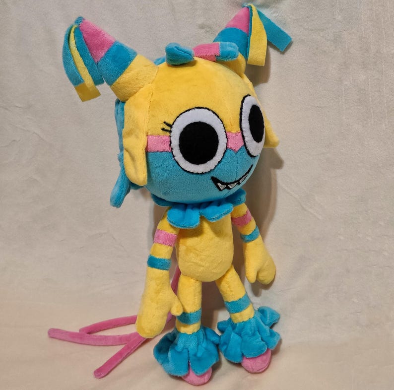 May include: A plush toy featuring a yellow head and body with bright blue and pink accents. The character has large white eyes with black pupils, a wide grin with visible teeth, and two tall, floppy ears. It wears blue ruffled cuffs and leg warmers.