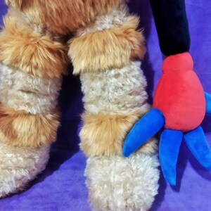 Peut inclure: Une peluche avec des jambes hirsutes de couleur cr&egrave;me et des accents de fourrure brune. Le jouet a un bras noir et une main rouge avec des appendices bleus en forme de doigts. Le fond est violet uni.