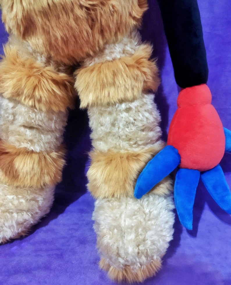 Peut inclure: Une peluche avec des jambes hirsutes de couleur cr&egrave;me et des accents de fourrure brune. Le jouet a un bras noir et une main rouge avec des appendices bleus en forme de doigts. Le fond est violet uni.