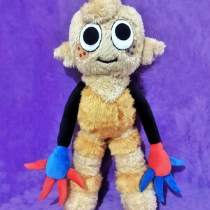 Peut inclure: Une peluche repr&eacute;sentant une cr&eacute;ature avec un corps brun clair et poilu, de grands yeux noirs et blancs et de petites oreilles. Elle a des bras noirs et des mains bleues et rouges. Le jouet est sur un fond violet.