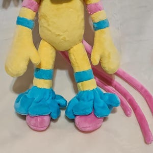May include: A plush toy featuring a yellow torso and limbs with bright blue bands on the arms and legs. The toy has blue ruffled cuffs at the ankles and pink rounded feet. Three long, thin pink appendages extend from the back of the plush figure.