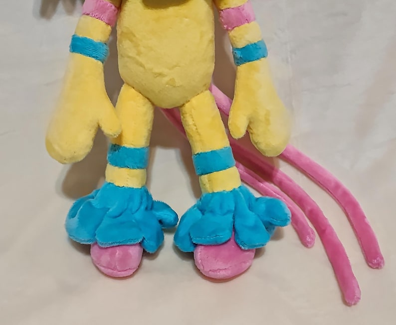 May include: A plush toy featuring a yellow torso and limbs with bright blue bands on the arms and legs. The toy has blue ruffled cuffs at the ankles and pink rounded feet. Three long, thin pink appendages extend from the back of the plush figure.
