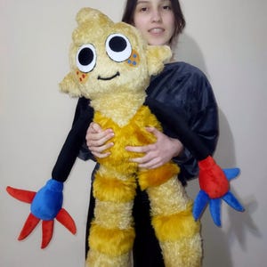 Peut inclure: Une grande peluche en forme de cr&eacute;ature de dessin anim&eacute;. Le jouet est principalement jaune avec des bras noirs et des appendices en forme de mains rouges et bleues. Il a de grands yeux ronds et une bouche souriante.