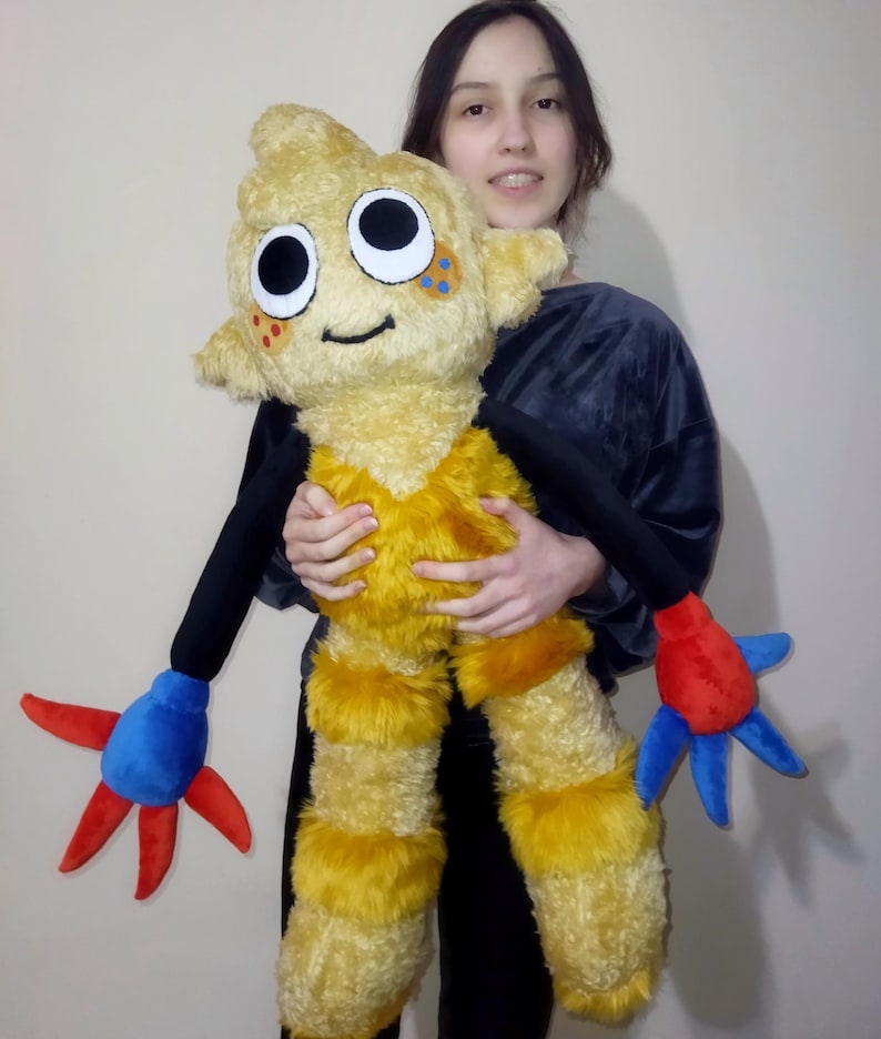 Peut inclure: Une grande peluche en forme de cr&eacute;ature de dessin anim&eacute;. Le jouet est principalement jaune avec des bras noirs et des appendices en forme de mains rouges et bleues. Il a de grands yeux ronds et une bouche souriante.