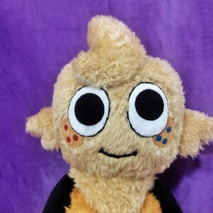 Peut inclure: Une peluche avec un ext&eacute;rieur brun clair et flou et de grands yeux noirs ronds. Le jouet a de petites oreilles et une touffe de cheveux sur le dessus. Il a une bouche souriante et des taches de points rouges et bleus sur les joues. Le fond est violet uni.