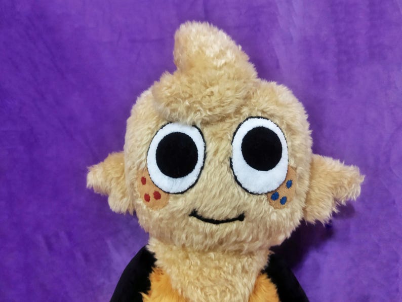 Peut inclure: Une peluche avec un ext&eacute;rieur brun clair et flou et de grands yeux noirs ronds. Le jouet a de petites oreilles et une touffe de cheveux sur le dessus. Il a une bouche souriante et des taches de points rouges et bleus sur les joues. Le fond est violet uni.
