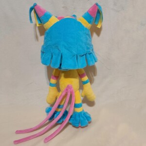 May include: A plush toy featuring a bright blue headpiece with pink and yellow accents, a yellow body with striped arms and legs, and three long pink tails. The character has blue ruffled feet and hands, with a soft texture throughout the design.