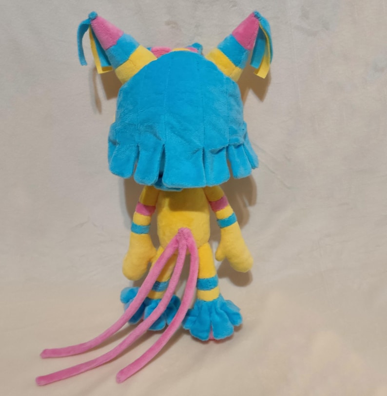 May include: A plush toy featuring a bright blue headpiece with pink and yellow accents, a yellow body with striped arms and legs, and three long pink tails. The character has blue ruffled feet and hands, with a soft texture throughout the design.