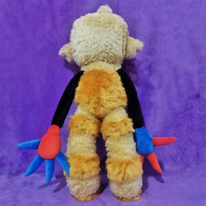 Peut inclure: Un singe en peluche avec une t&ecirc;te et un corps brun clair, des bras noirs et de la fourrure orange sur la poitrine et les jambes. Les mains sont bleues et rouges. Le jouet est sur un fond violet.