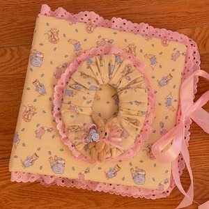 May include: A square fabric-covered photo album featuring a light yellow background with a repeating pattern of small illustrated animals. The album is edged with pink eyelet lace and includes a central pleated fabric oval with a small plush bunny.