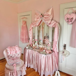 May include: A pink vanity set with a large satin bow and a mirror. The vanity has a white and pink skirt and is decorated with lace. There is a pink chair with a white lace skirt and a pink cushion. The vanity is set in a pink room with white trim.