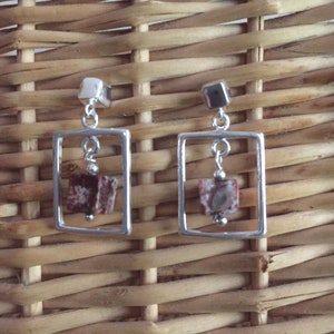 May include: A pair of silver earrings with a square frame and a red and white stone hanging inside. The earrings are on a woven wicker background.