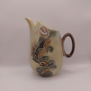 May include: A ceramic pitcher with a light green glaze and brown accents. The pitcher features a raised design of stylized flowers and leaves.