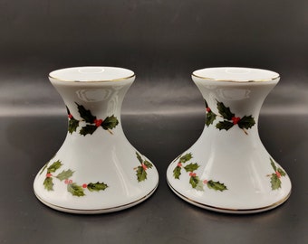 May include: Two white ceramic candle holders with a gold rim and a holly berry design. The candle holders have a flared base and a narrow neck. The holly design features green leaves and red berries, perfect for holiday decor.