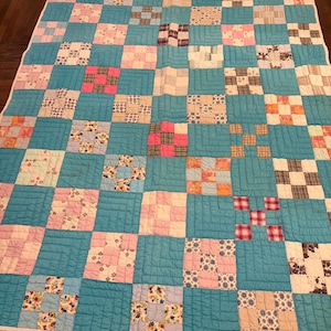 May include: A patchwork quilt featuring a grid of solid turquoise squares alternating with multi-colored patterned squares in shades of pink, white, tan, and floral prints. The quilt has visible machine-stitched quilting lines throughout.