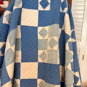 May include: A quilt featuring a geometric patchwork pattern in shades of medium blue, light blue, and off-white. The design consists of alternating squares and diamond shapes with an overall quilted texture visible across the fabric surface.